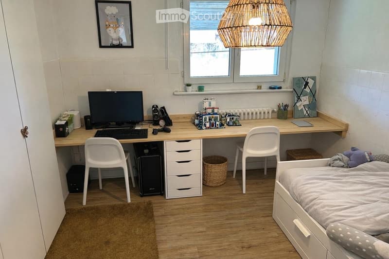 A room with a desk, two chairs, a bed, a lamp, a picture frame, a window, and a radiator.