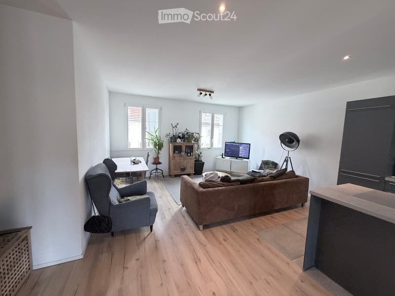 Living room with wooden floor, white walls, brown couch, wooden table, TV, potted plants, lamp, and a chair