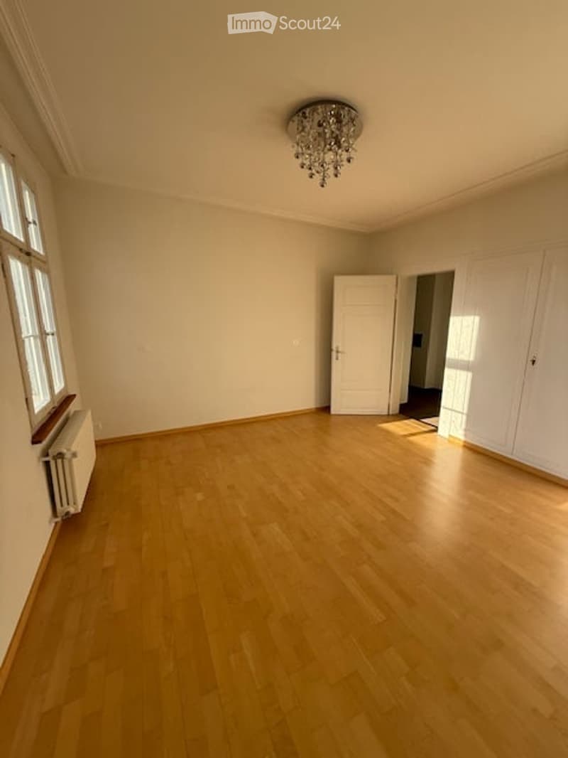 Empty room with wooden floor, white walls, large windows, chandelier, radiator, two doors