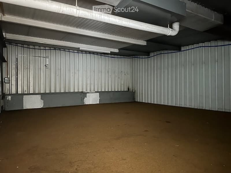 Empty room with white walls, brown floor, metal ceiling, pipes, and a power outlet.
