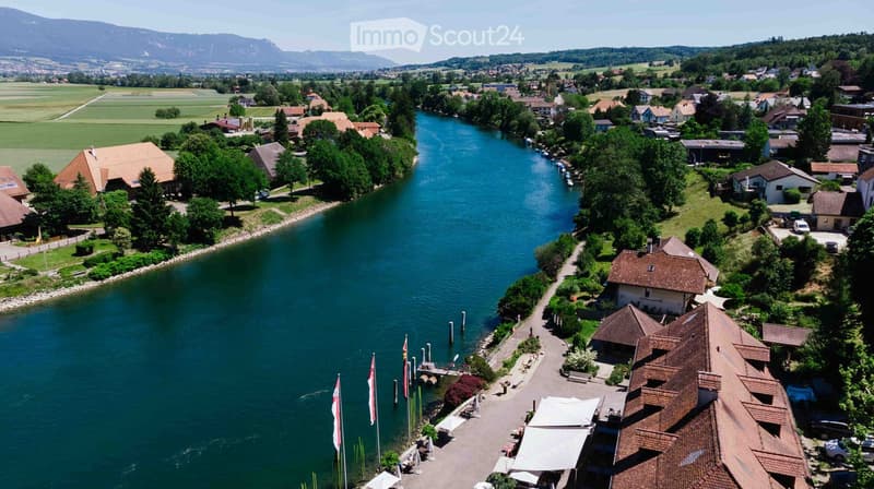 Aerial view of a town with a river running through it, surrounded by green fields and mountains in the background. Houses and buildings are visible along the riverbanks.