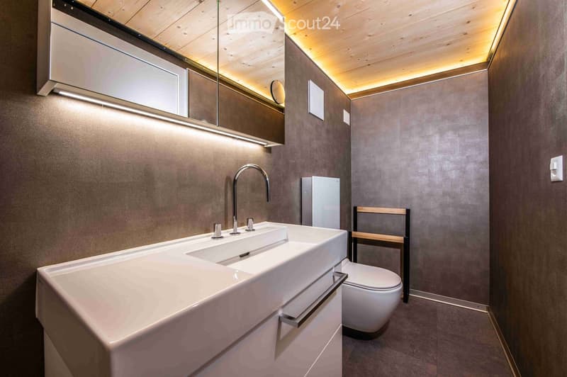 Modern bathroom with a double sink, sleek faucet, large mirror, toilet, and a small wooden stool. The walls are textured grey, and the ceiling is wooden with integrated lighting.