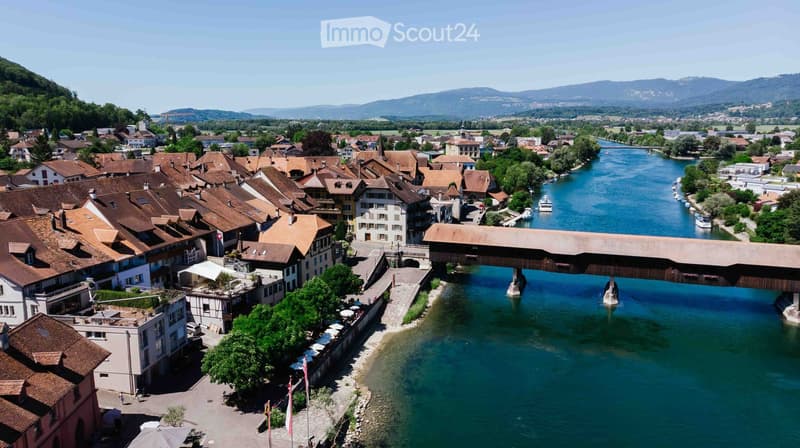 Aerial view of a town with traditional houses, a wooden bridge over a river, and surrounding mountains.