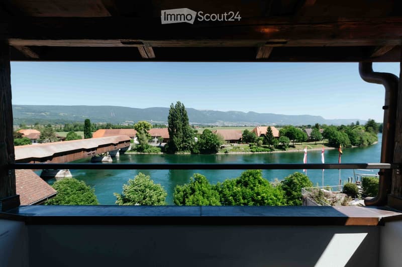 Balcony overlooking a scenic river valley with a wooden covered bridge, distant mountains, and surrounding greenery.