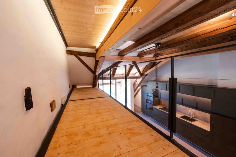 Modern hallway with wooden flooring, exposed wooden beams, and a glass partition leading to a kitchen area with stainless steel appliances and dark cabinets.