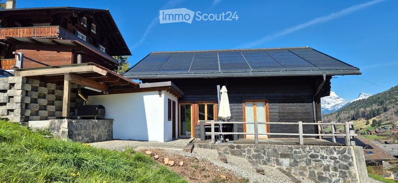 Detached house, wooden exterior, solar panels on the roof, stone wall foundation, balcony, terrace, outdoor seating, mountain view