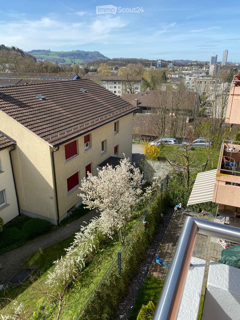 Aerial view of a house with a brown roof, red shutters, and a balcony with a view of the city and mountains in the distance.