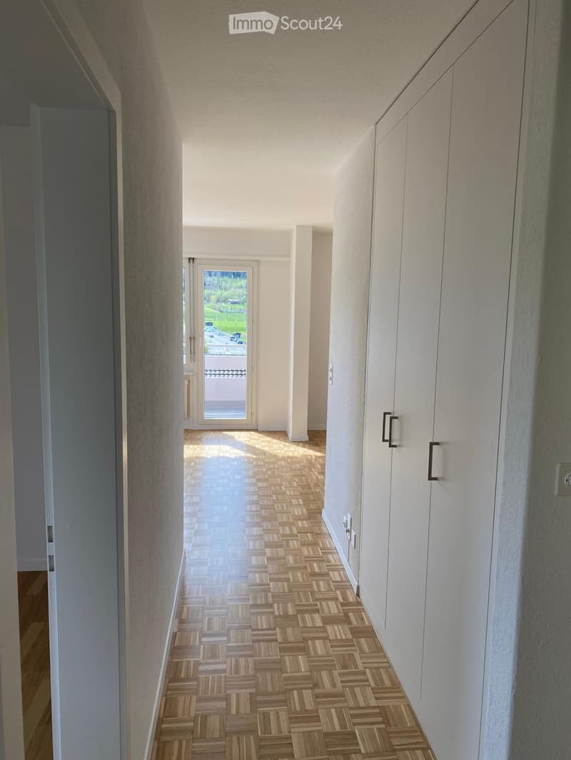 Empty hallway with wooden parquet flooring, white walls, sliding glass door leading to a balcony, and built-in wardrobes on the right side.