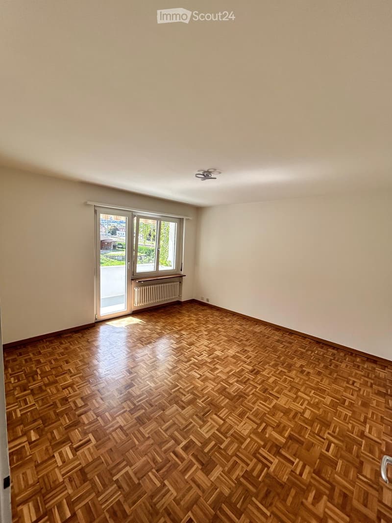 Empty room with wooden parquet flooring, glass sliding doors leading to a balcony, white walls, white ceiling, radiator, ceiling fan, window with white frames.