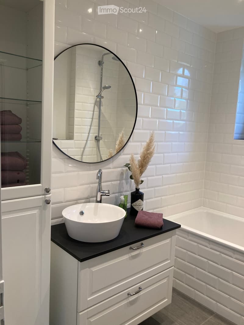 white tiles, round mirror, white sink, black countertop, white bathtub, white cabinets, glass cabinet