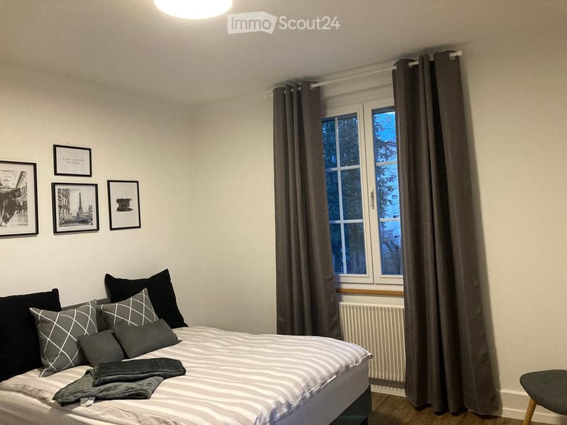 Bedroom with bed, pillows, blankets, radiator, window with curtains, framed pictures on wall