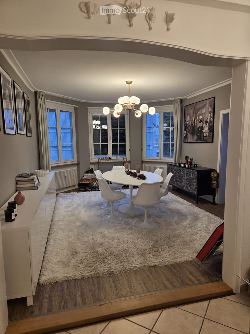Dining room with white table and chairs, chandelier, white rug, white cabinets, white radiator, picture frames, black cabinet, books, and decorative items