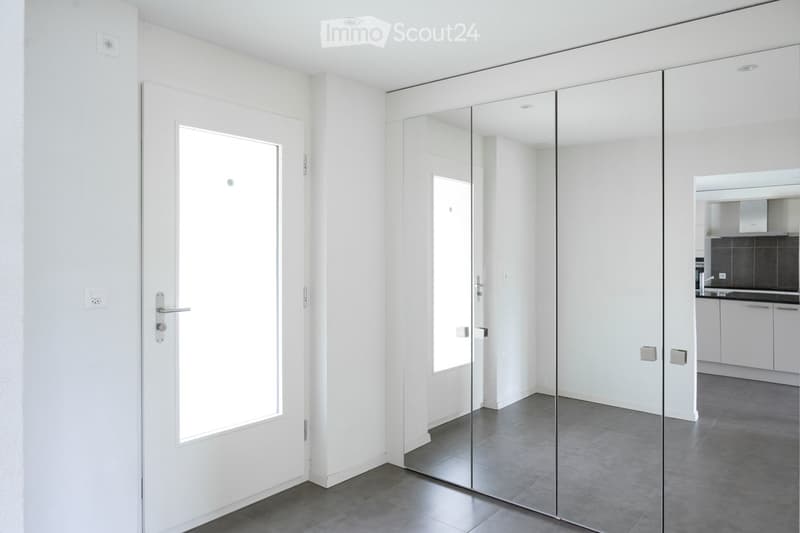 Corridor / Hallway. Modern, minimalist hallway with white walls, grey tiled floor, large mirrored doors, and a white door leading to another room.