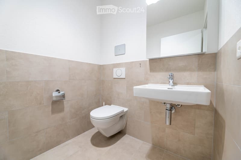 Bathroom with white toilet, white sink, mirror, beige tiles, and silver fixtures