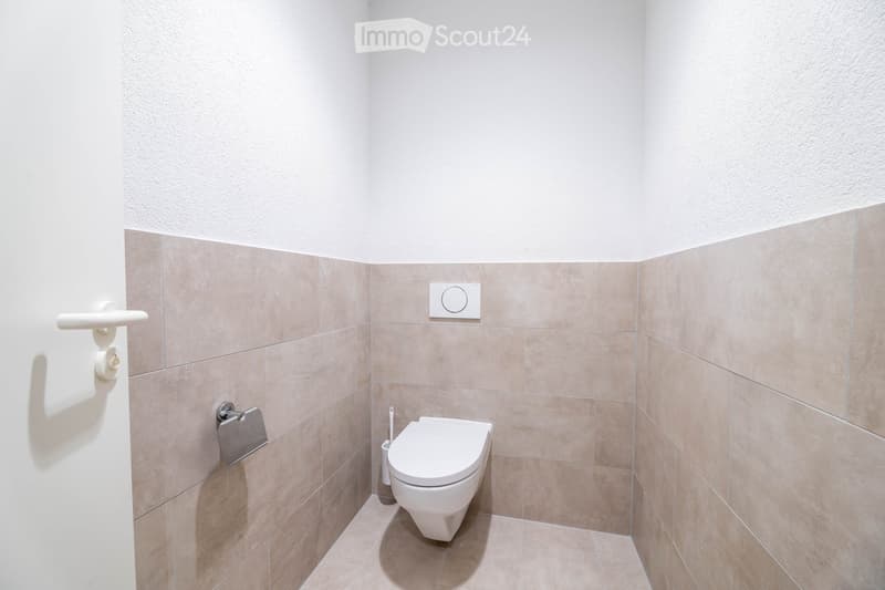 small bathroom with white toilet, beige tiles, white walls, toilet paper holder, toilet brush