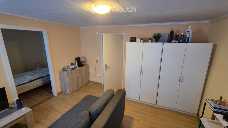Living room with couch, wooden floor, white walls, ceiling light, cabinet, bed, TV, and plants