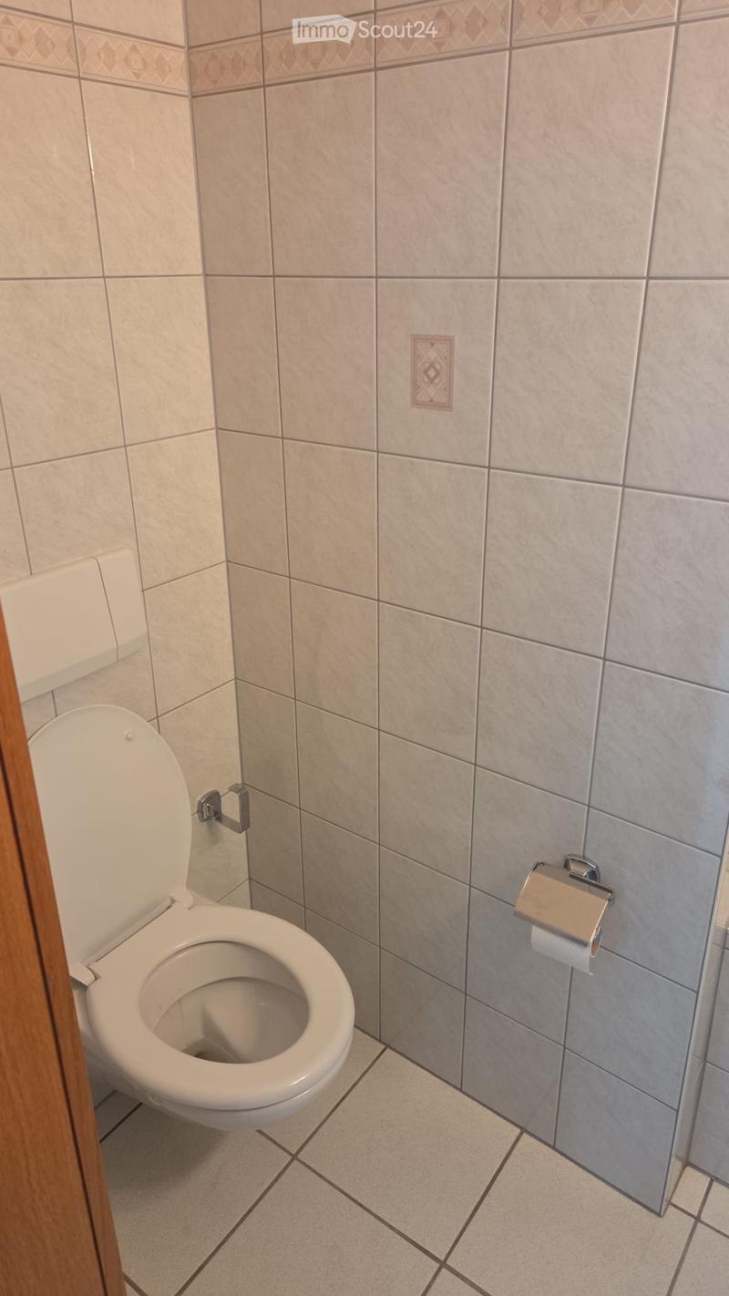 A small bathroom with white tiles, a toilet, and a toilet paper holder on the wall.
