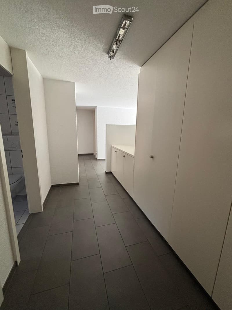 A narrow hallway with tiled flooring, white walls, and built-in storage cabinets on one side. The hallway leads to a bathroom with a visible toilet and shower area.