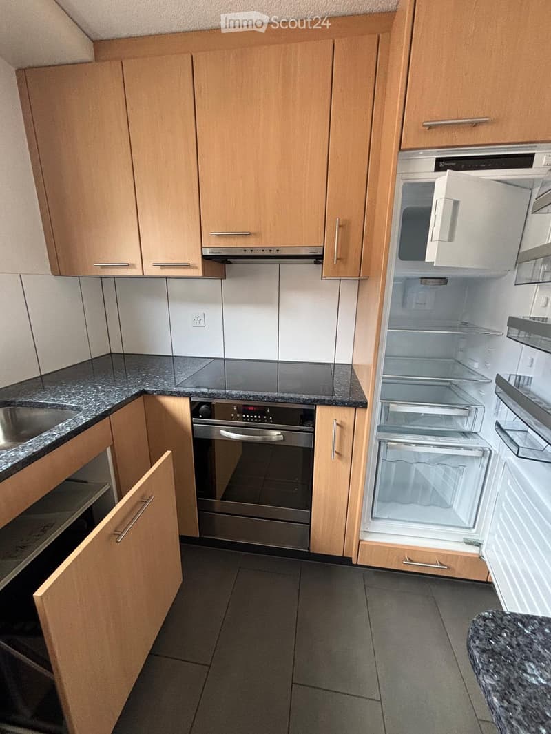 Modern kitchen with light wooden cabinets, granite countertops, stainless steel appliances including a refrigerator, oven, and microwave. The refrigerator door is open, revealing its interior compartments.