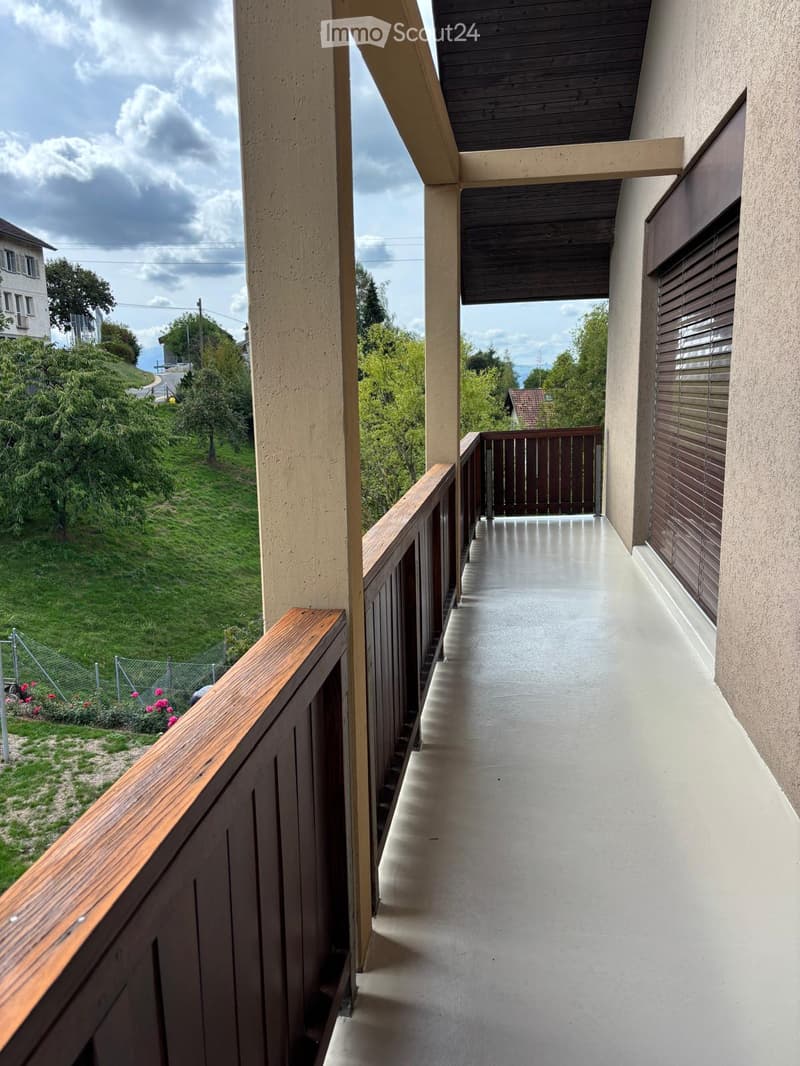 Balcony with wooden railing, concrete floor, and a view of a green hillside and neighboring houses.