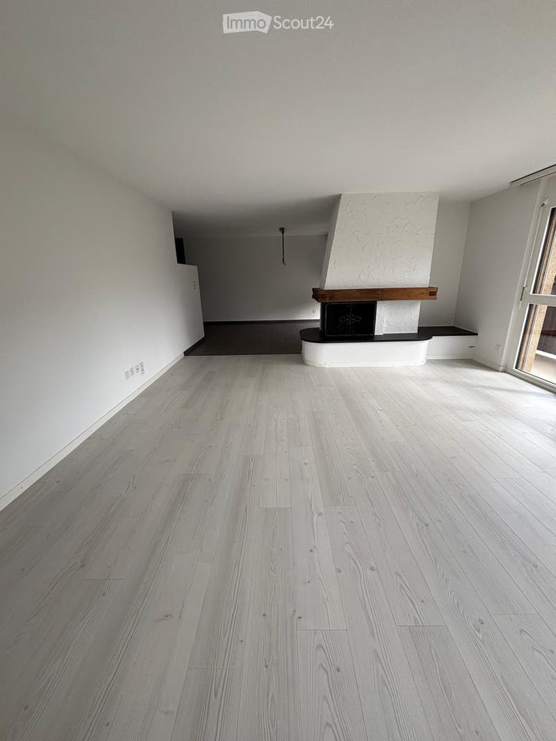 Empty room with light grey wooden flooring, white walls, and a fireplace with a black insert and wooden mantelpiece.