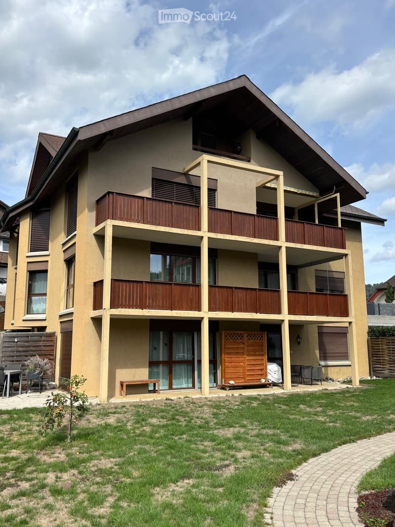 Two-story house with beige facade, wooden balconies, and a well-maintained lawn in front.