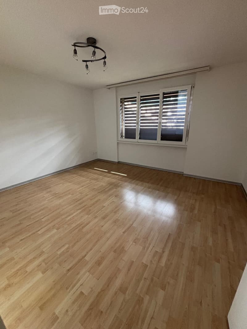 Empty room with wooden flooring, white walls, and a large window with blinds. A modern chandelier hangs from the ceiling.