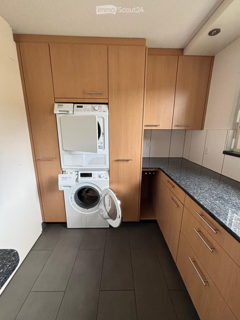 A small, well-lit room with a washing machine and wooden cabinets, featuring a dark tiled floor and a grey countertop.