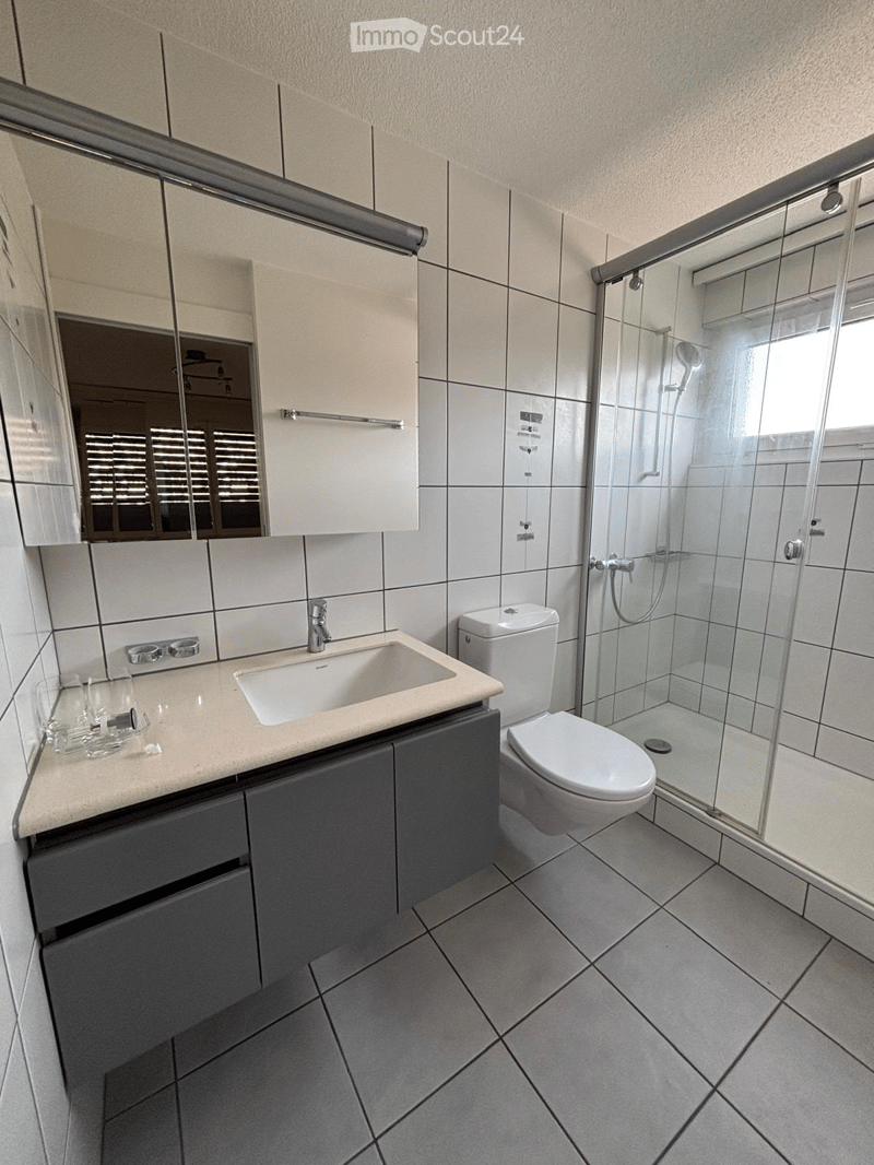 Modern bathroom with white tiles, a sink with a mirror, a toilet, and a glass-enclosed shower. The countertop has a beige finish, and the floor is tiled in gray.