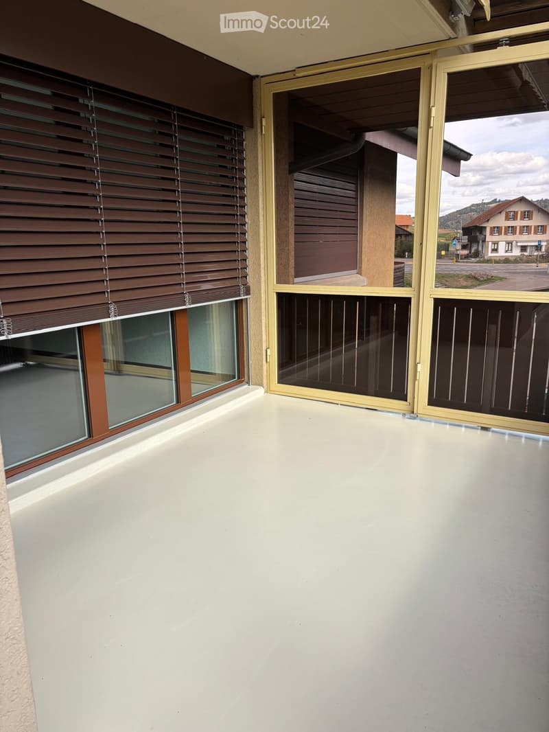 Balcony with white floor, large sliding glass doors, wooden blinds on one side, and a view of a street and houses.