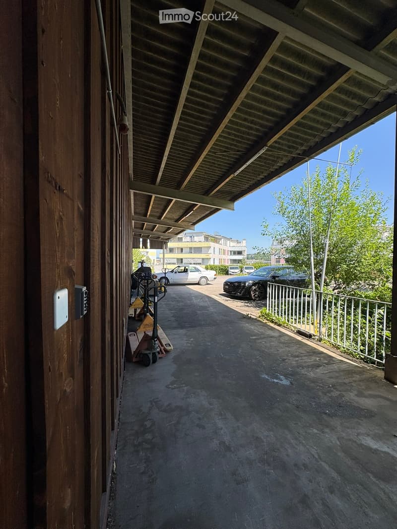 A covered outdoor corridor with a concrete floor, metal railing on one side, and several parked cars visible in the background.