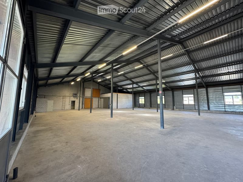 Large, empty industrial-style space with a concrete floor, metal ceiling, and exposed structural beams. The room has large windows on one side and a few doors leading to other areas.