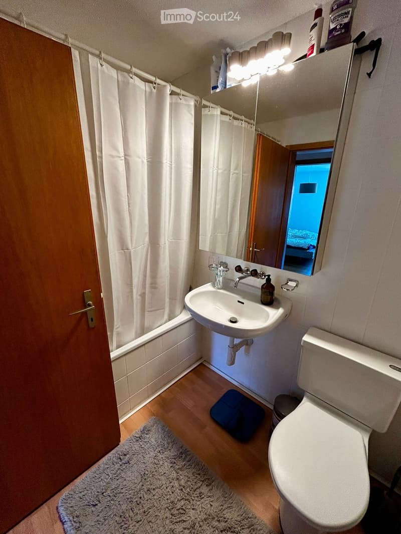 Bathroom with wooden floor, white walls, white bathtub, white sink, mirror, toilet, rug, and white shower curtain