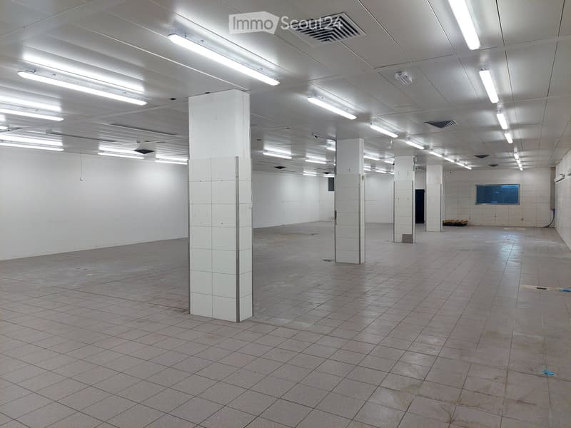 Empty room with white tiled floor, white walls, and ceiling with multiple fluorescent lights, several white pillars, and a window.