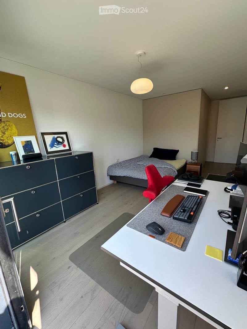A room with a bed, desk, and cabinet. The bed has a striped blanket and pillows. The desk has a keyboard, mouse, and monitor. The cabinet has picture frames and a cup. The room has a wooden floor and a white ceiling with a lamp.