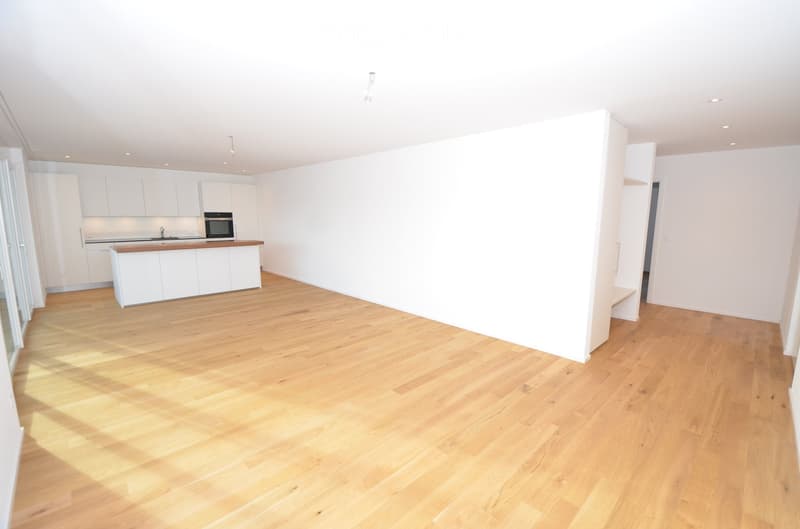 Empty room with wooden floor, white walls, kitchen with island, white cabinets, built-in oven, sink, and faucet