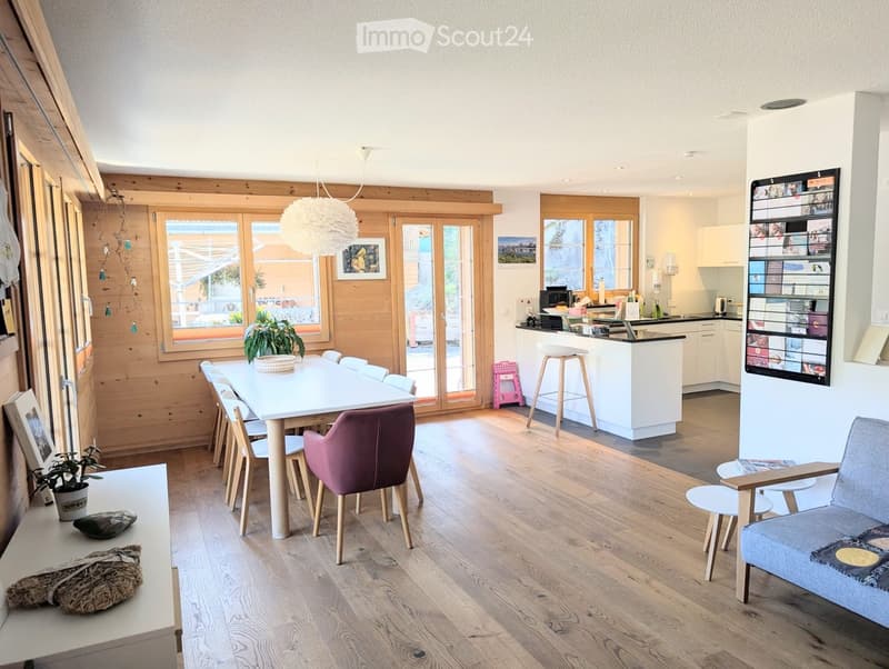 Dining area with a large table, wooden chairs, wooden floor, wooden wall, kitchen with a counter, white cabinets, and a small table with a stool