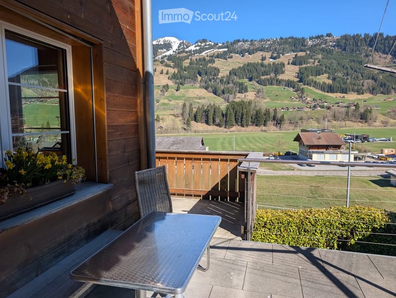 Balcony with table and chair, wooden wall, window with flowers, metal railing, view of mountains and fields