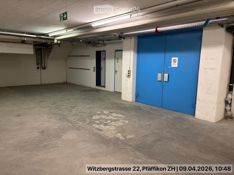 Empty parking garage with concrete floor, white walls, blue doors, and metal pipes on the ceiling.