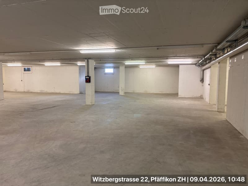 Empty parking lot, concrete floor, white walls, fluorescent lights, small windows, central pillar