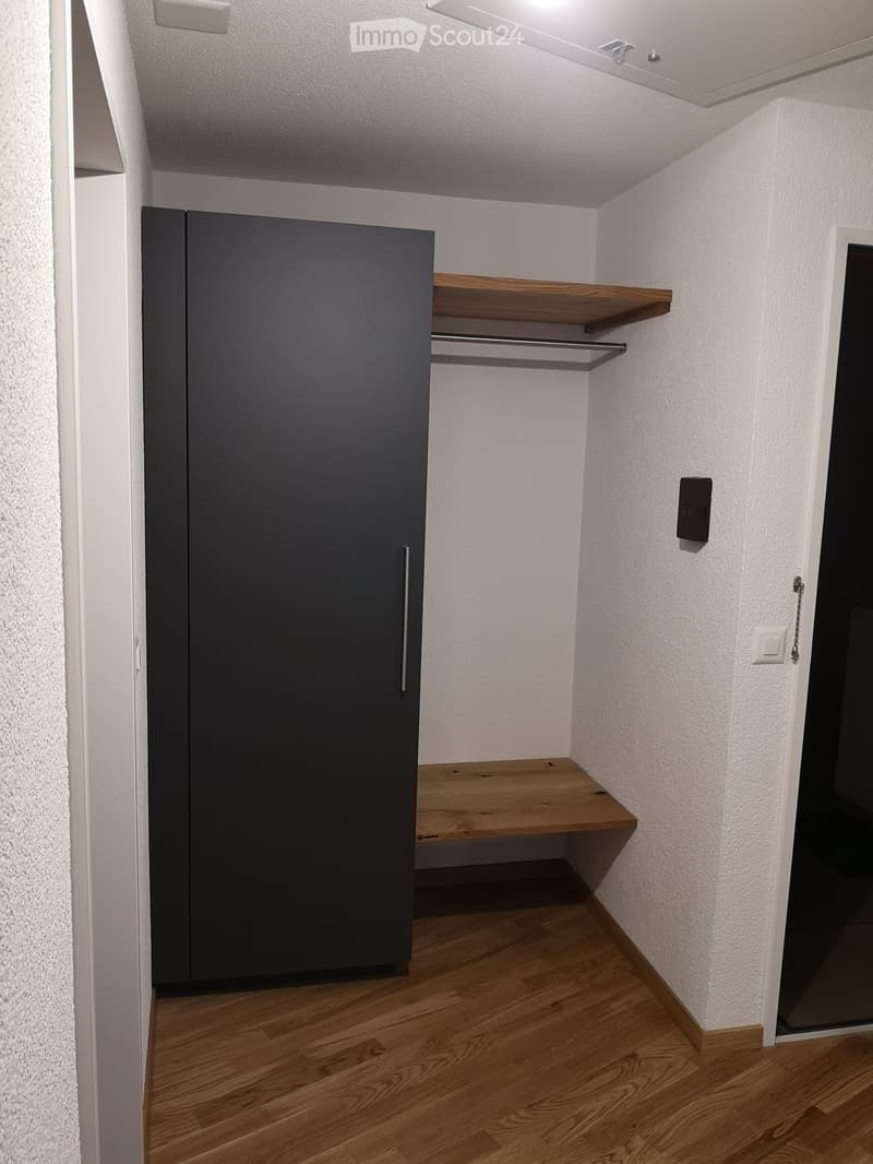 A small room with a black wardrobe, wooden shelf, and a white wall.