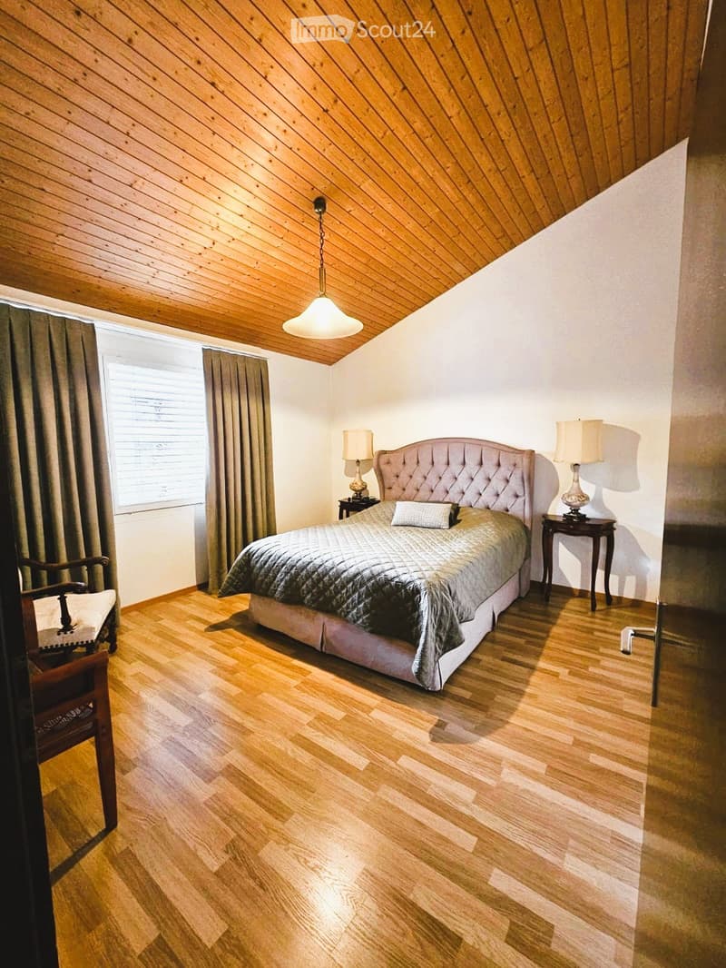 Bedroom with wooden floor, wooden ceiling, bed, nightstands, lamps, chair, window with curtains