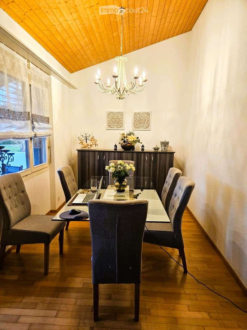 Dining room with wooden floor, chandelier, dining table, chairs, flower vase, laptop, and a cabinet with decorative items