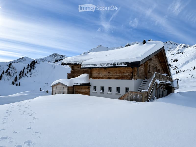A wooden chalet with snow-covered roof and surrounding snowy landscape, featuring a staircase leading to a balcony.