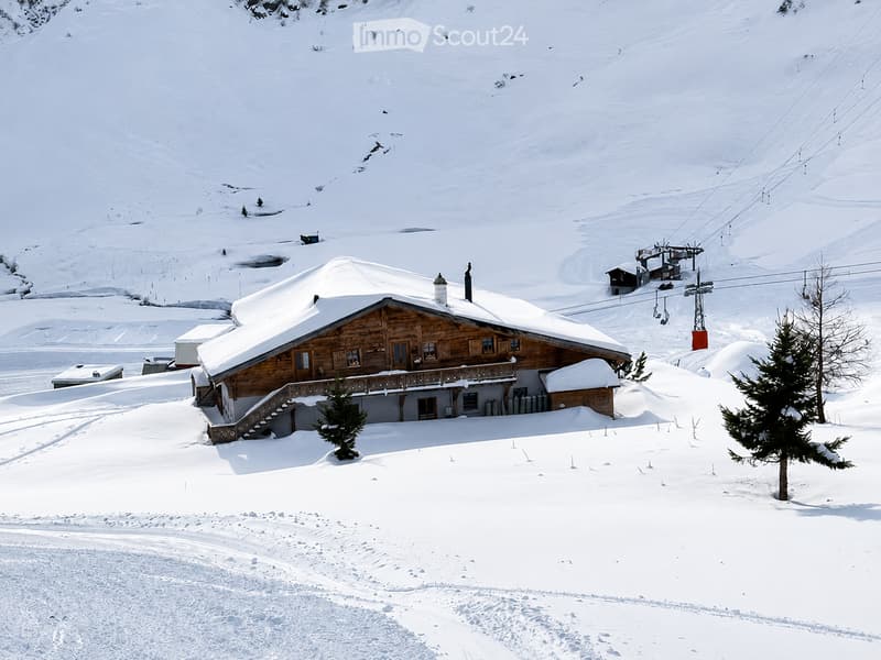 A wooden house with a snow-covered roof and balcony, situated in a snowy mountain landscape with ski lift cables visible in the background.