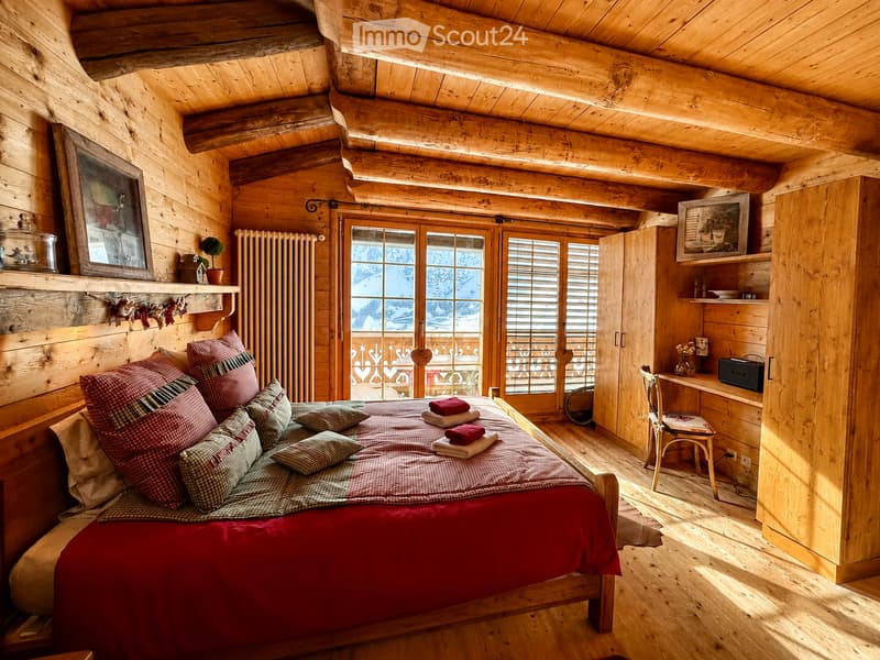 Cozy wooden bedroom with a large bed, colorful pillows, a wooden chair, a small table, shelves, and a framed picture on the wall. Large windows offer a scenic view.