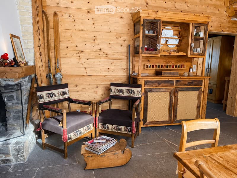 Cozy living room with wooden walls, a stone fireplace, two patterned armchairs, a wooden table, a cabinet with glass doors, and a small stool holding magazines.