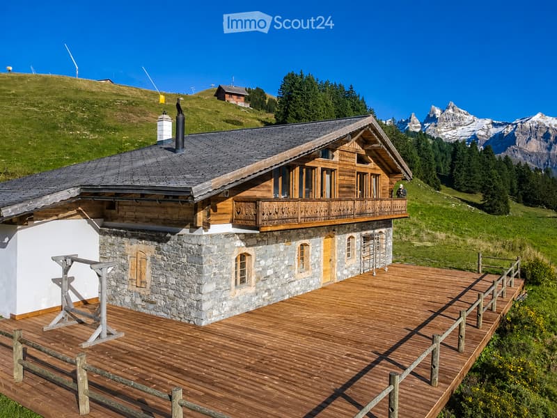 A two-story wooden chalet with stone base, large balcony, and wooden deck, surrounded by green hills and mountains.