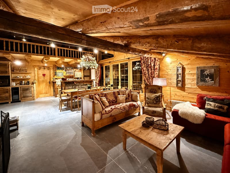 Cozy living room with wooden walls and ceiling, featuring a sofa, coffee table, armchairs, dining area with table and chairs, and a small bar area.
