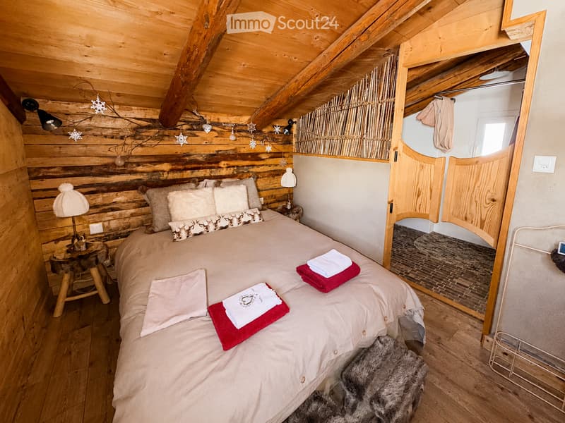 Cozy attic bedroom with wooden beams, a large bed with pillows and blankets, a nightstand with a lamp, and a fur rug on the floor. A small bathroom is visible through an open wooden door.